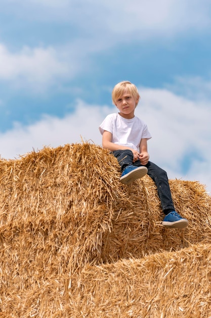 Blonder Junge mit europäischem Aussehen in Jeans und T-Shirt sitzt auf Heuhaufen vor blauem Himmelshintergrund