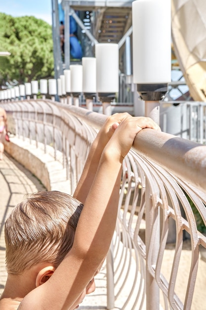 Blonder Junge mit nassen Haaren hält sich an einem sonnigen Tag am Geländer in der Nähe der großen Rutsche im Wasserpark fest