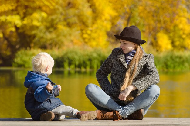 Blonder Junge mit seiner Mutter, die auf dem Pier sitzt und mit Äpfeln spielt. Sonniger Herbsttag