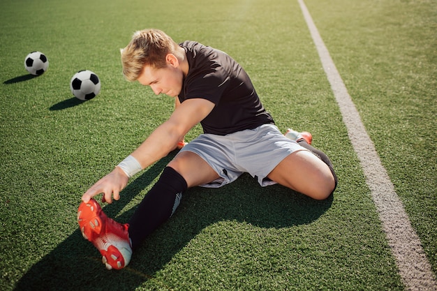 Blonder junger Fußballspieler strecken sich draußen auf Rasen. Er erreicht die Füße mit der Hand. Guy konzentriert sich auf Bewegung. Zwei Fußbälle liegen hinter ihm.