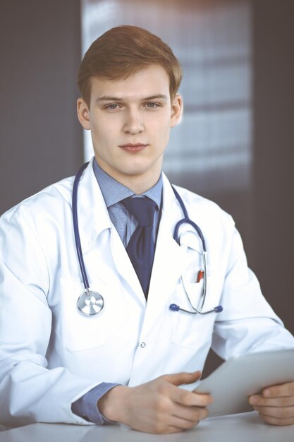 Foto blonder männlicher arzt sitzt und arbeitet mit tablet-computer in der sonnigen klinik an seinem arbeitsplatz, nahaufnahme. medizinkonzept