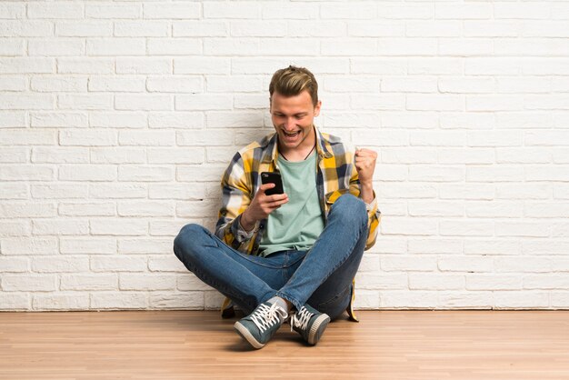 Blonder Mann, der auf dem Boden mit Telefon in Siegposition sitzt