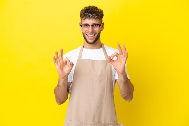 Blonder Mann des Restaurantkellners lokalisiert auf gelbem Hintergrund, der ein okayzeichen mit den Fingern zeigt