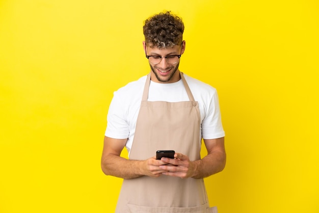 Blonder Mann des Restaurantkellners lokalisiert auf gelbem Hintergrund, der eine Nachricht mit dem Handy sendet