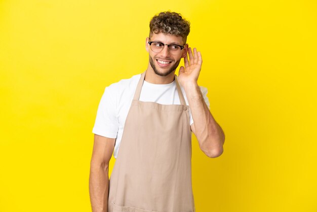 Blonder Mann des Restaurantkellners lokalisiert auf gelbem Hintergrund, der etwas hört, indem er Hand auf das Ohr legt