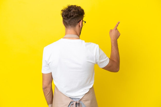 Blonder Mann des Restaurantkellners lokalisiert auf gelbem Hintergrund, der mit dem Zeigefinger zurück zeigt