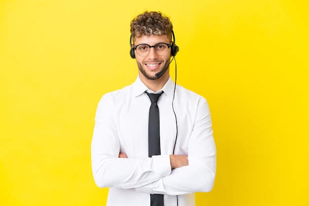 Blonder Mann des Telemarketers, der mit einem Headset arbeitet, das auf gelbem Hintergrund isoliert ist und die Arme in Frontalposition verschränkt hält