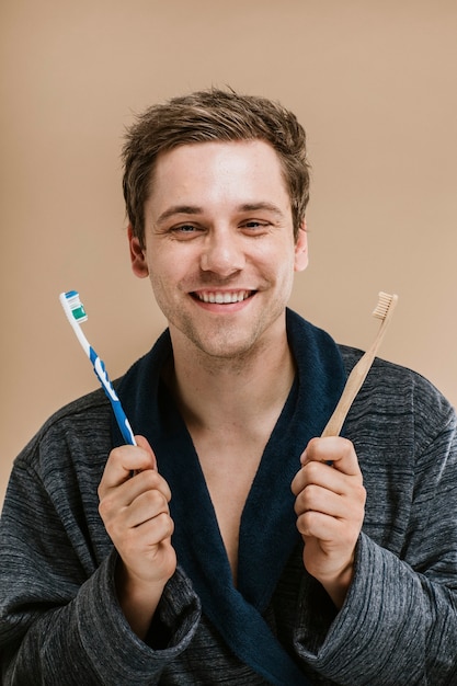 Blonder mann in einem gewand, der zwischen einer holzzahnbürste und einer plastikzahnbürste wählt