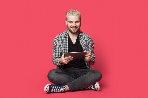 Blonder Mann mit Bart sitzt auf dem Boden mit einem Tablet an einer roten Wand