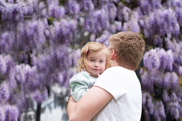 Blonder Mann mit süßer Tochter, die in die Kamera blickt, in der Nähe von Glucinum-Blumen, lila, sehr peri wistar