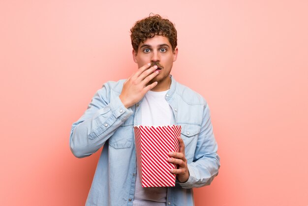 Blonder Mann über der rosa Wand überrascht und Popcorn essend