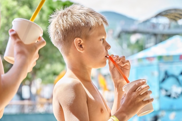 Blonder Schuljunge trinkt leckeres Getränk mit Strohhalm im Café im Freien des Wasserparks im Sommerresort