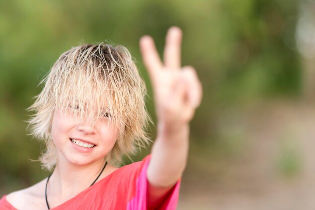 Foto blonder teenager des mädchens in der roten bluse, der handgeste sieg zeigt