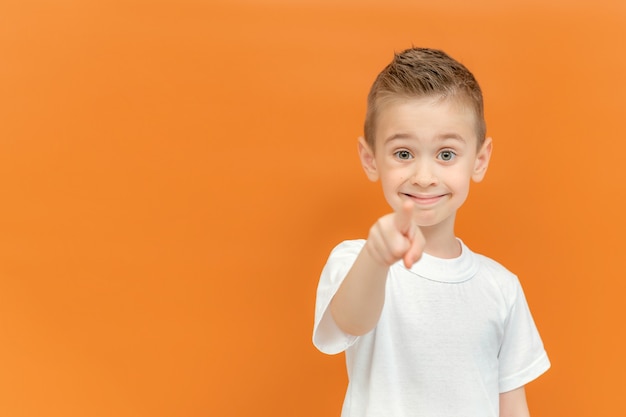 Blonder Vorschuljunge, der auf Kamera mit glücklichem und lustigem Gesicht zeigt, das auf orange Hintergrund lokalisiert wird. Hübsches energisches kleines kaukasisches Kind, das weißes T-Shirt trägt. Platz kopieren.