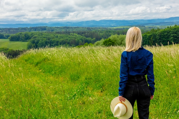 Blondes Cowgirl im Hut auf Wiese mit Bergen