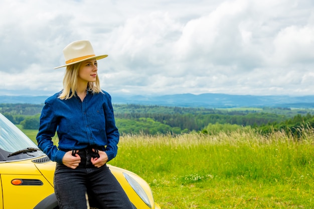 Blondes Cowgirl im Hut nahe einem Auto auf Wiese mit Bergen