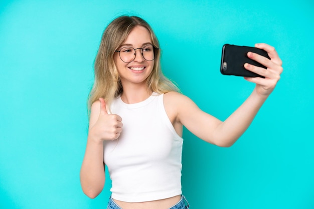 Blondes englisches junges Mädchen lokalisiert auf blauem Hintergrund, der ein selfie mit Handy macht