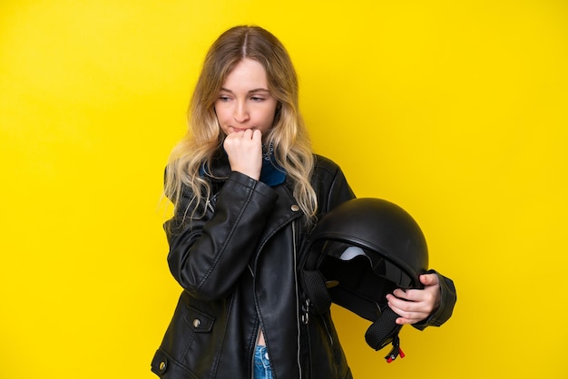 Blondes englisches junges Mädchen mit einem Motorradhelm, der auf gelbem Hintergrund isoliert ist und Zweifel hat