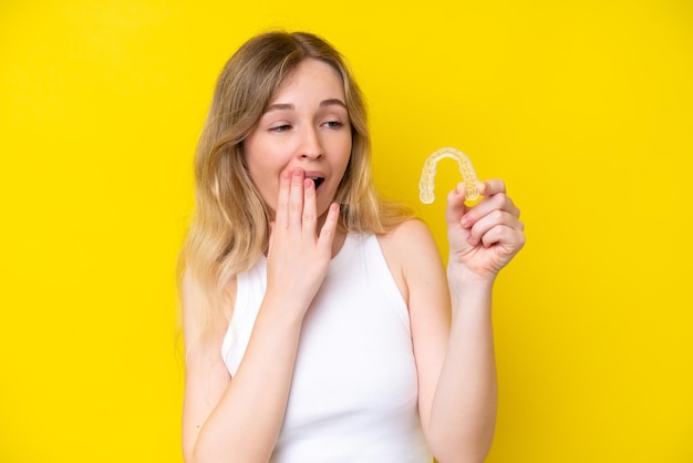 Blondes englisches junges Mädchen mit unsichtbaren Zahnspangen isoliert auf gelbem Hintergrund mit überraschtem und schockiertem Gesichtsausdruck