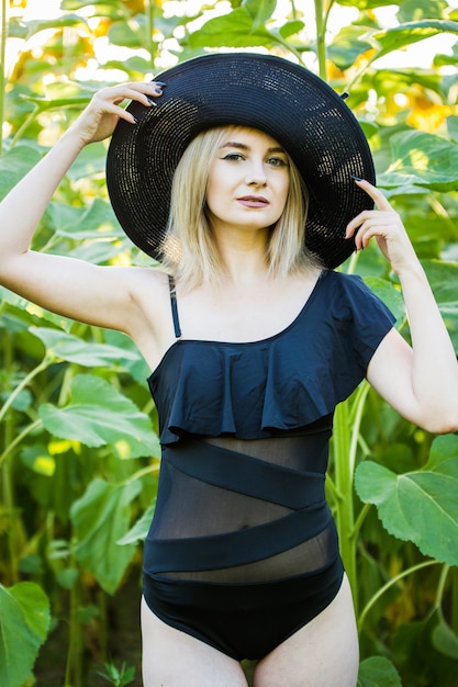 Blondes europäisches Mädchen in einem schwarzen Badeanzug und Hut auf Natur mit Sonnenblumen