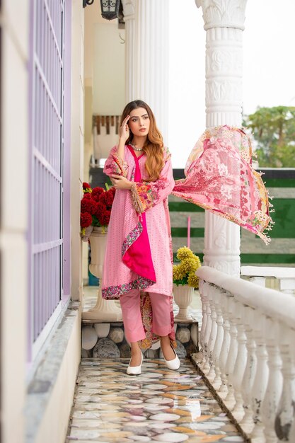 Blondes Haarmädchen posiert mit winkendem Dupatta auf Terrasse mit Blumen, die Designer Shalwar Kameez tragen