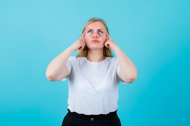 Blondes junges Mädchen schaut nach oben, indem es den Zeigefinger auf Schläfen auf blauem Hintergrund hält