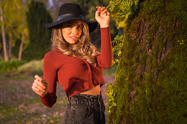 Blondes kaukasisches Lebensstil-Mädchen in einem roten Pullover und in einem schwarzen Hut, die Natur in einem Park mit Baumporträt der attraktiven jungen Frau im Herbst genießen