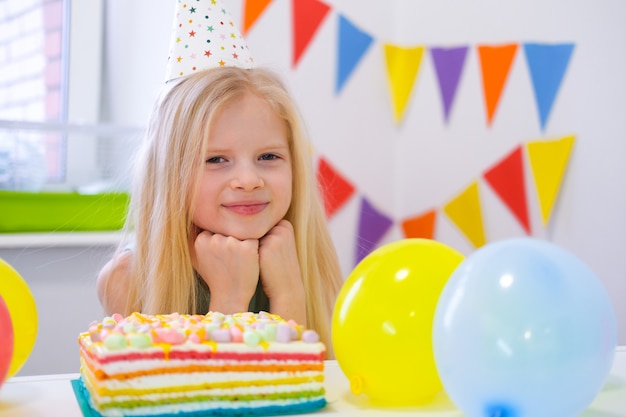 Blondes kaukasisches Mädchen sitzt nachdenklich und träumerisch am festlichen Tisch nahe Geburtstagsregenbogenkuchen und macht einen Wunsch. Kamera betrachten. Bunter Hintergrund mit Ballonen