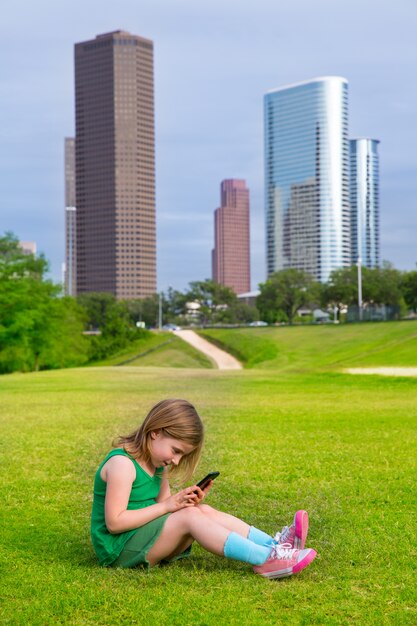 Blondes Kindermädchen, das mit dem Smartphone sitzt auf Parkrasen an den Stadtskylinen spielt