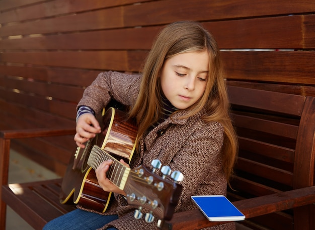 Blondes Kindermädchen, das Spielgitarre mit Smartphone lernt
