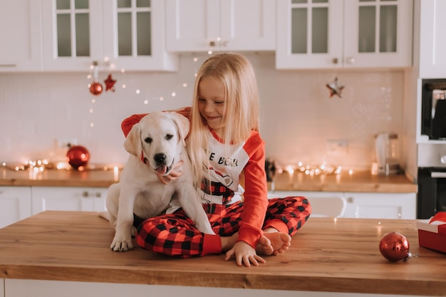 Blondes kleines Mädchen im roten Weihnachtspyjama sitzt mit einem weißen Hund in einer bekränzten Küche. Winterwochenenden und Feiertage im herzlichen Familienkreis. Kind und Haustier. Platz für Text. Foto in hoher Qualität