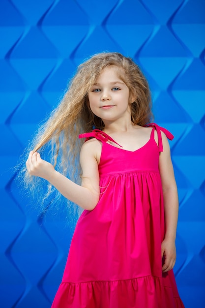 Blondes kleines Mädchen in einem rosa Kleid auf blauem Hintergrund tanzen und lachen, helle Kindergefühle der Freude, glückliche Kindheit
