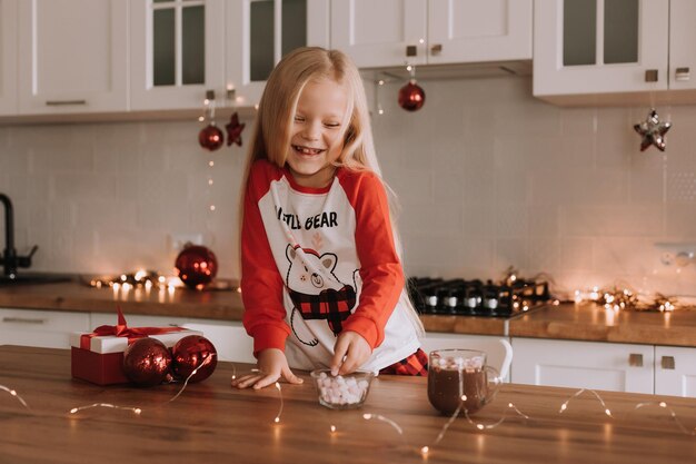 Blondes kleines Mädchen in rotem Weihnachtspyjama fügt Marshmallows zu einem Becher mit einem heißen Getränk in einer wunderschön dekorierten Küche für den Urlaub hinzu. Lebensstil. Platz für Text. Foto in hoher Qualität