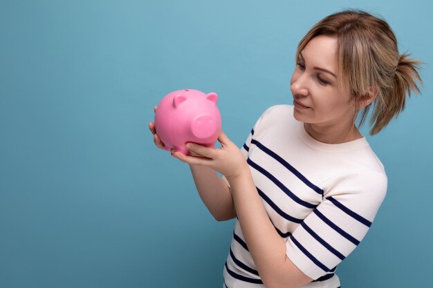 Blondes lächelndes Mädchen im legeren Outfit, das ein Sparschwein auf blauem Hintergrund mit Kopierraum hält