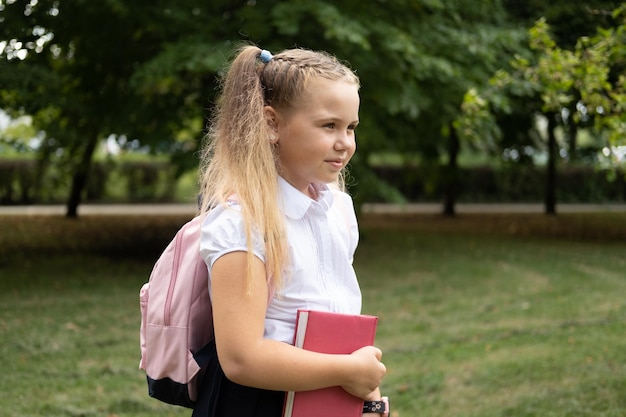 blondes lächelndes schulmädchen in schuluniform, das notizbuch mit rosa rucksack zurück zur schule hält