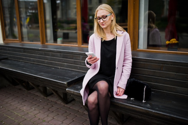 Blondes Mädchen an den Gläsern und am rosa Mantel, schwarzer Kittel, der auf Bank an der Straße sitzt und auf Smartphone schaut.