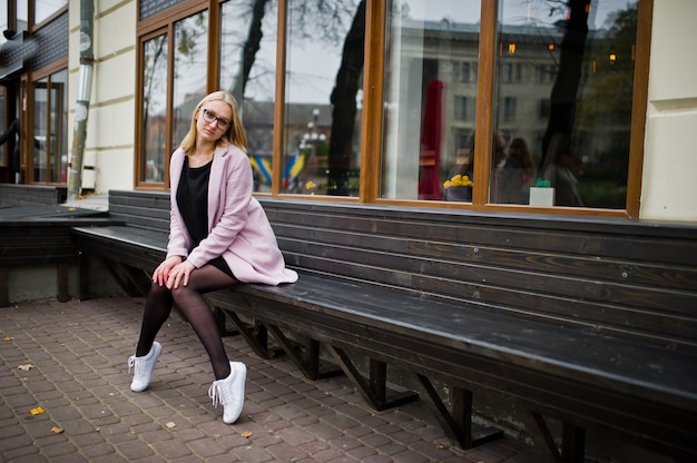 Blondes Mädchen an den Gläsern und am rosa Mantel, schwarzer Kittel, der auf Bank an der Straße sitzt.