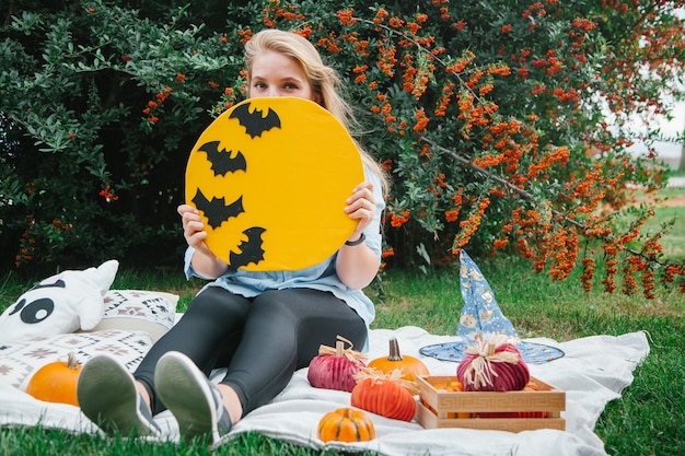 Blondes Mädchen auf der Decke im Park auf dem Rasen mit einem Geist und Kürbissen Halloween-Symbolen