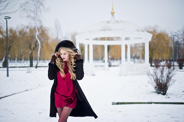 Blondes Mädchen der Eleganz im Pelzmantel und im roten Abendkleid stellte am verschneiten Wintertag auf