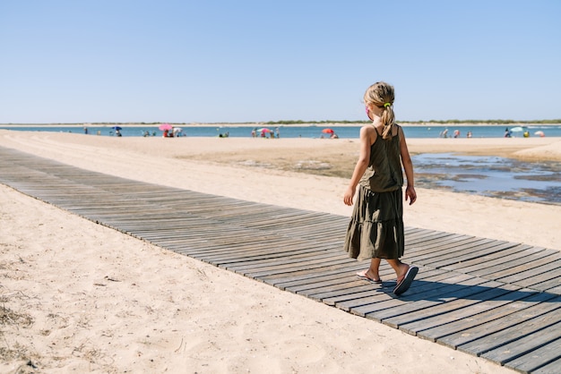 Blondes Mädchen der Rückansicht mit Gesichtsmaske im Urlaub, der durch einen Weg im Sand eines Strandes mit einem grünen Kleid mitten in einer Coronavirus-Pandemie geht