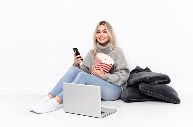 Blondes Mädchen des Jugendlichen, das Popcorn beim Aufpassen eines Films auf dem Laptop hält Kaffee zum Mitnehmen und ein Mobile isst