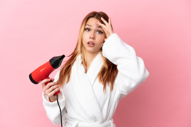 Blondes Mädchen des Teenagers, das einen Haartrockner über lokalisiertem rosa Hintergrund hält und Überraschungsgeste macht, während er zur Seite schaut