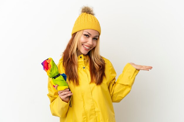 Blondes Mädchen des Teenagers, das einen regendichten Mantel über lokalisiertem weißem Hintergrund trägt, der Kopienraum imaginär auf der Handfläche hält, um eine Anzeige einzufügen