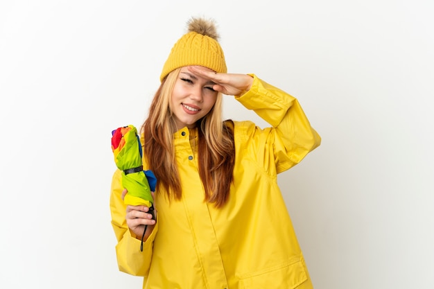 Blondes Mädchen des Teenagers, das einen regenfesten Mantel über lokalisiertem weißem Hintergrund trägt, der mit der Hand weit weg schaut, um etwas zu schauen