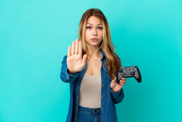 Blondes Mädchen des Teenagers, das mit einem Videospielcontroller über einer isolierten Wand spielt und eine Stoppgeste macht