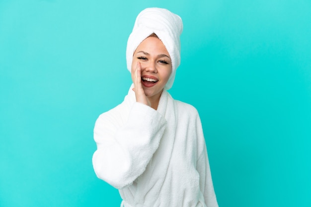 Blondes Mädchen des Teenagers in einem Bademantel über lokalisiertem blauem Hintergrund, der mit weit geöffnetem Mund schreit