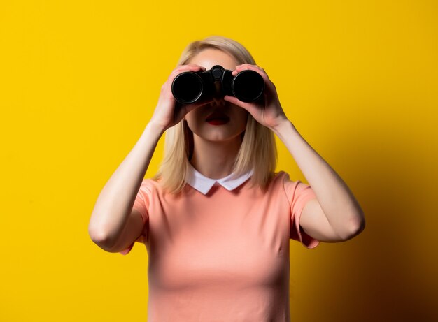 Blondes Mädchen im rosa Kleid, das im Fernglas auf gelbem Raum beobachtet