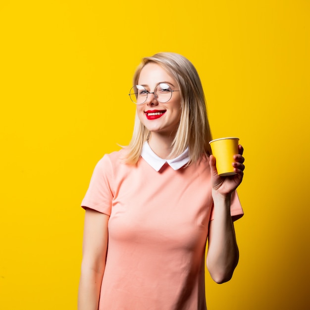 Blondes Mädchen im rosa Kleid mit Pappbecher auf gelbem Raum