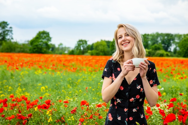 Blondes Mädchen im schönen Kleid mit Tasse Kaffee im Mohnfeld in der Sommerzeit