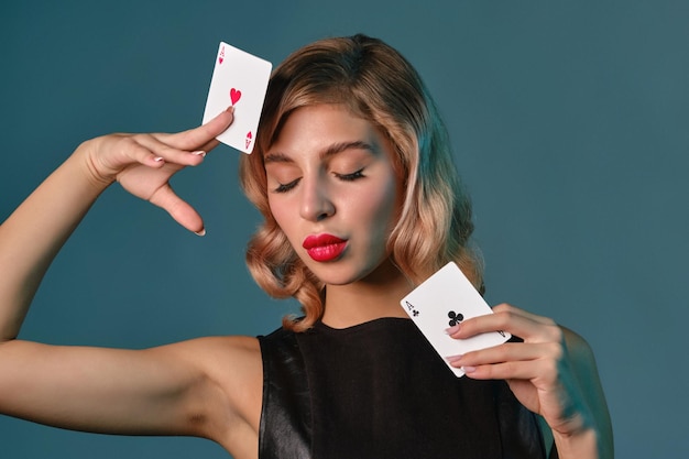 Blondes Mädchen im schwarzen Lederkleid, das zwei Spielkarten zeigt, die vor blauem Hintergrund posieren Glücksspiel Unterhaltung Poker Casino Closeup
